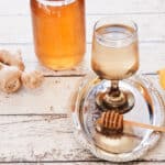 A glass of hot ginger tea with a bit of honey dipper covered with honey and ginger in the background.