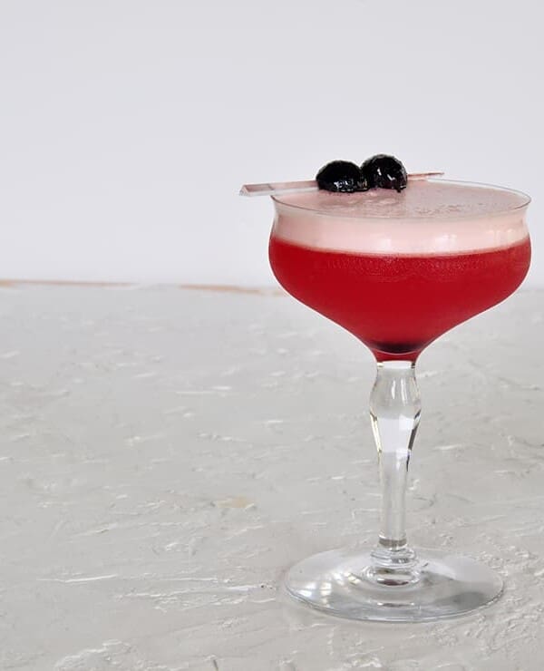 A Single Sam Ross Sour Cocktail on a white backtdrop and garnished with two maraschino cherries