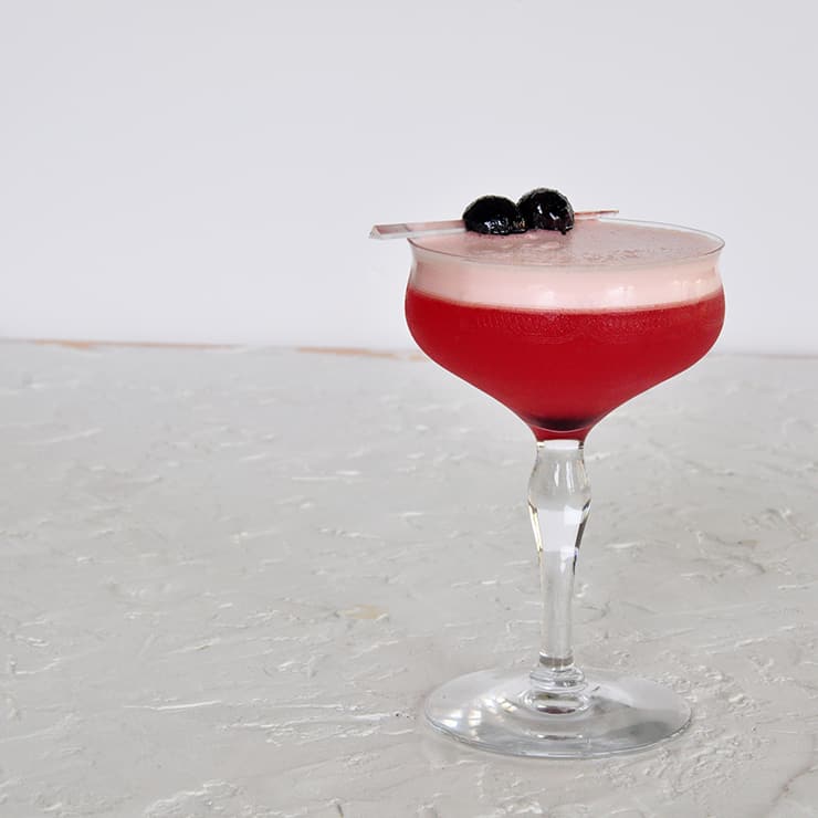 A Single Sam Ross Sour Cocktail on a white backtdrop and garnished with two maraschino cherries