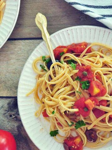 A plate of Puttanesca Sauce with Linguine on a white plate with a gold spoon. It is in a picnic table with a striped white cloth and some whole tomatoes.
