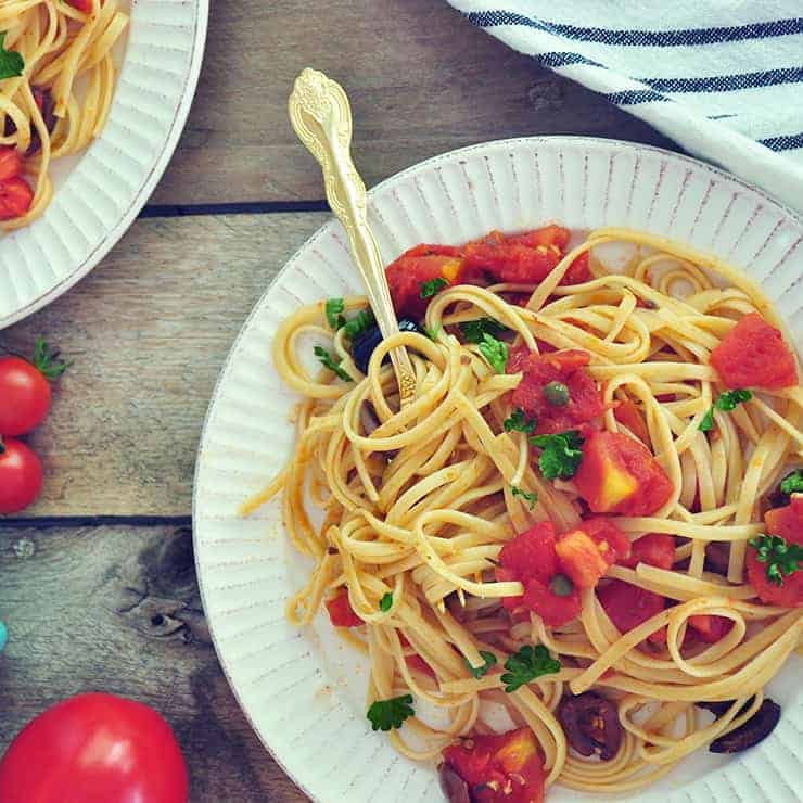 A plate of Puttanesca Sauce with Linguine on a white plate with a gold spoon. It is in a picnic table with a striped white cloth and some whole tomatoes.