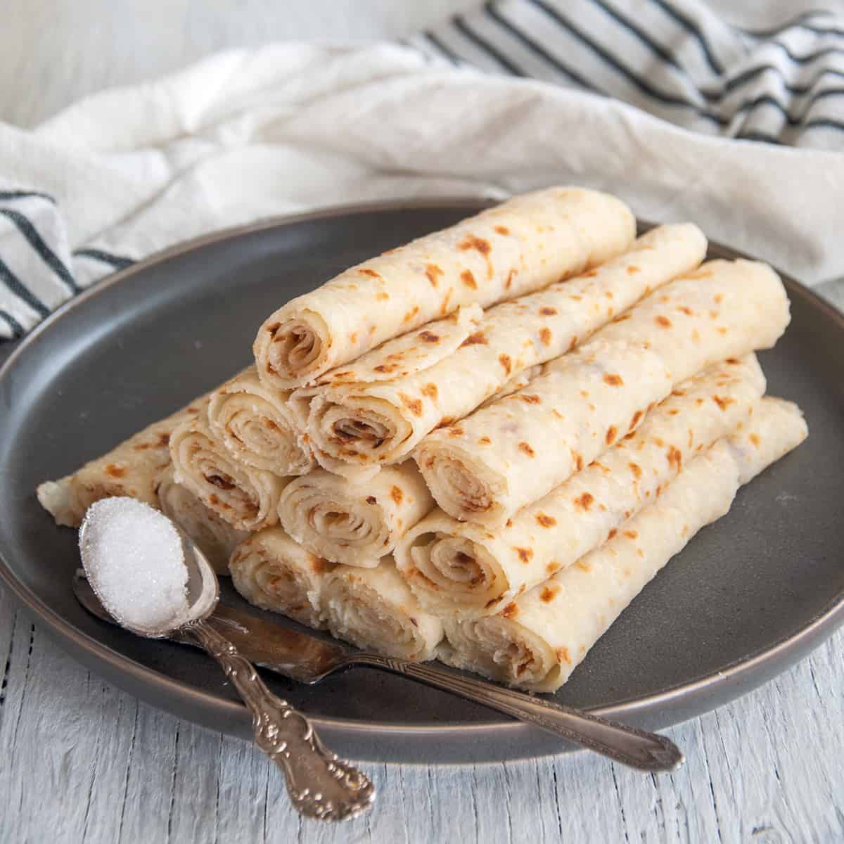 Pile of lefse from this lefse recipe on a dark plate with a knife and a spoonful of sugar.