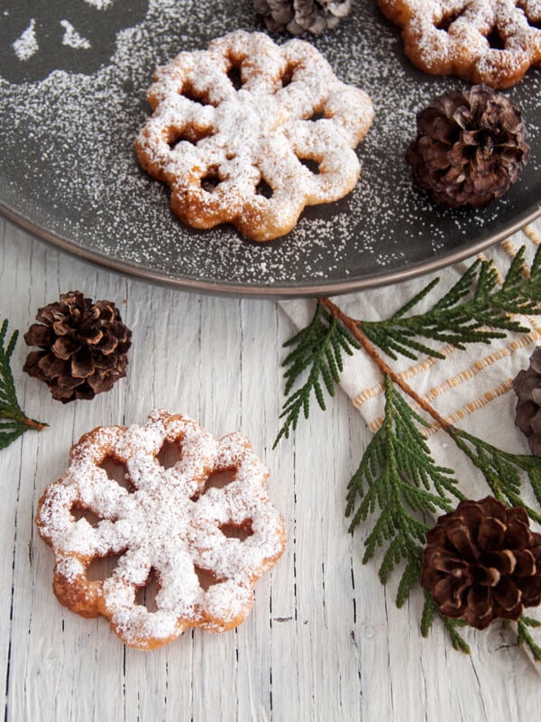 A rosette cookie on a white background with evergreen and pinecones around it and a few other ones on a plate behind it.