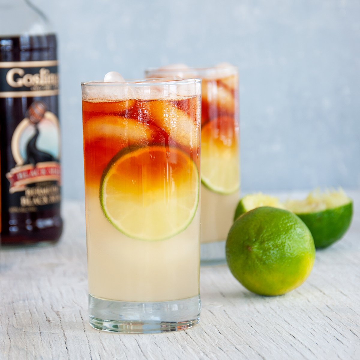 Two Dark 'n Stormy cocktails on a white table with limes and a bottle of Gosling's dark rum in the background.