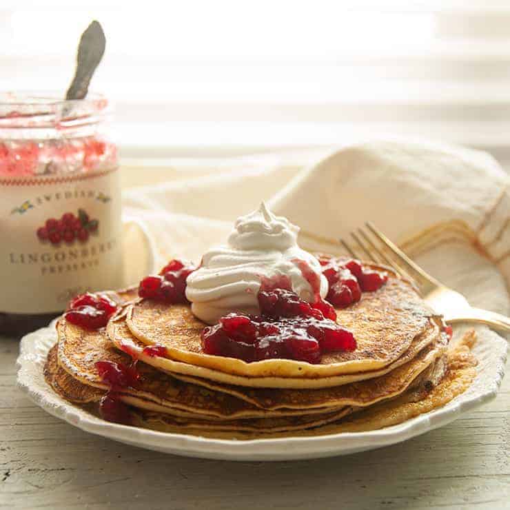 Stack of Swedish Pancakes Recipe on a white plate with Lingonberry jam, whipped cream, and powdered sugar on top.