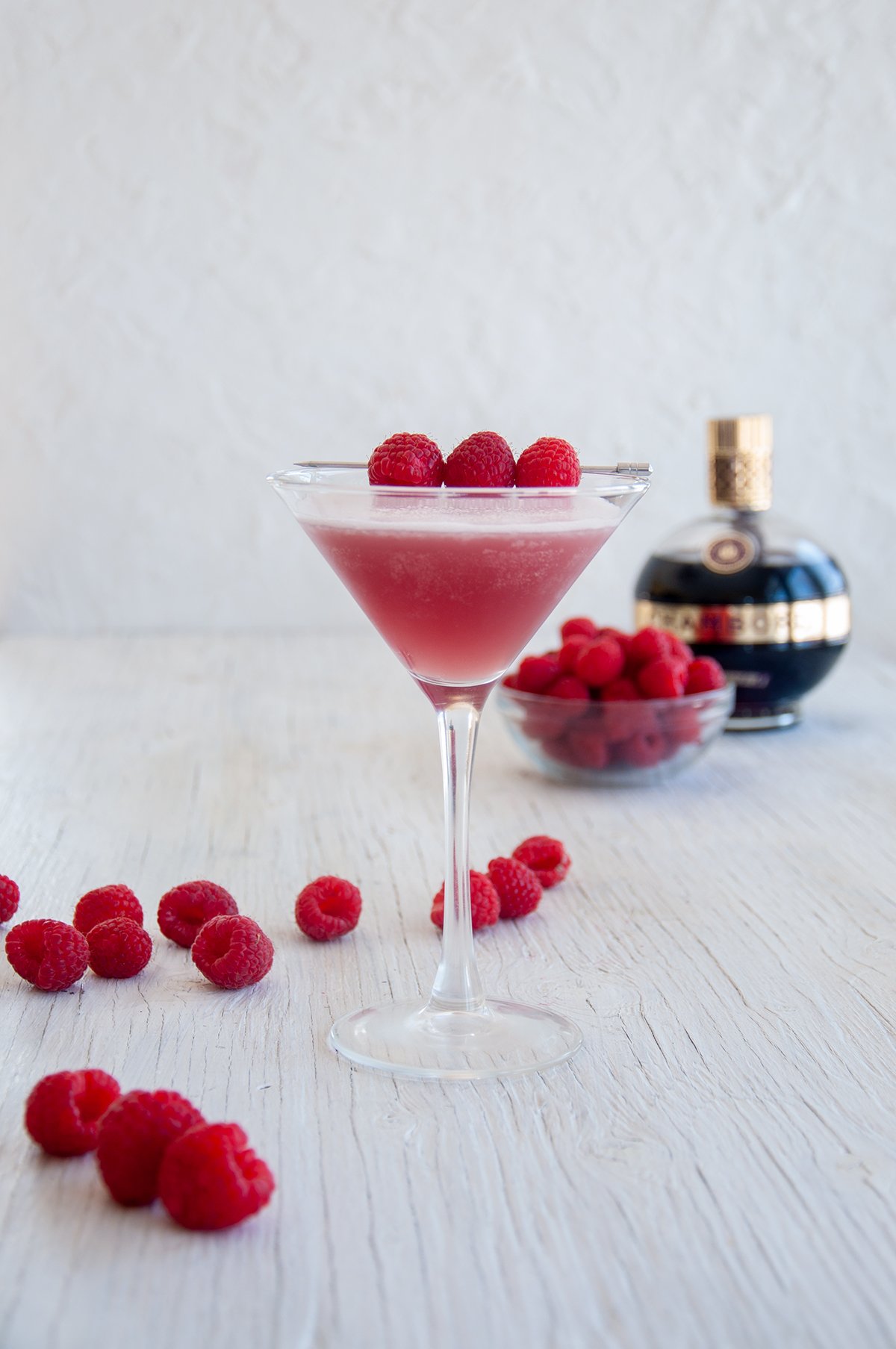 One French Martini on a white table with raspberries and Chambord in the background.
