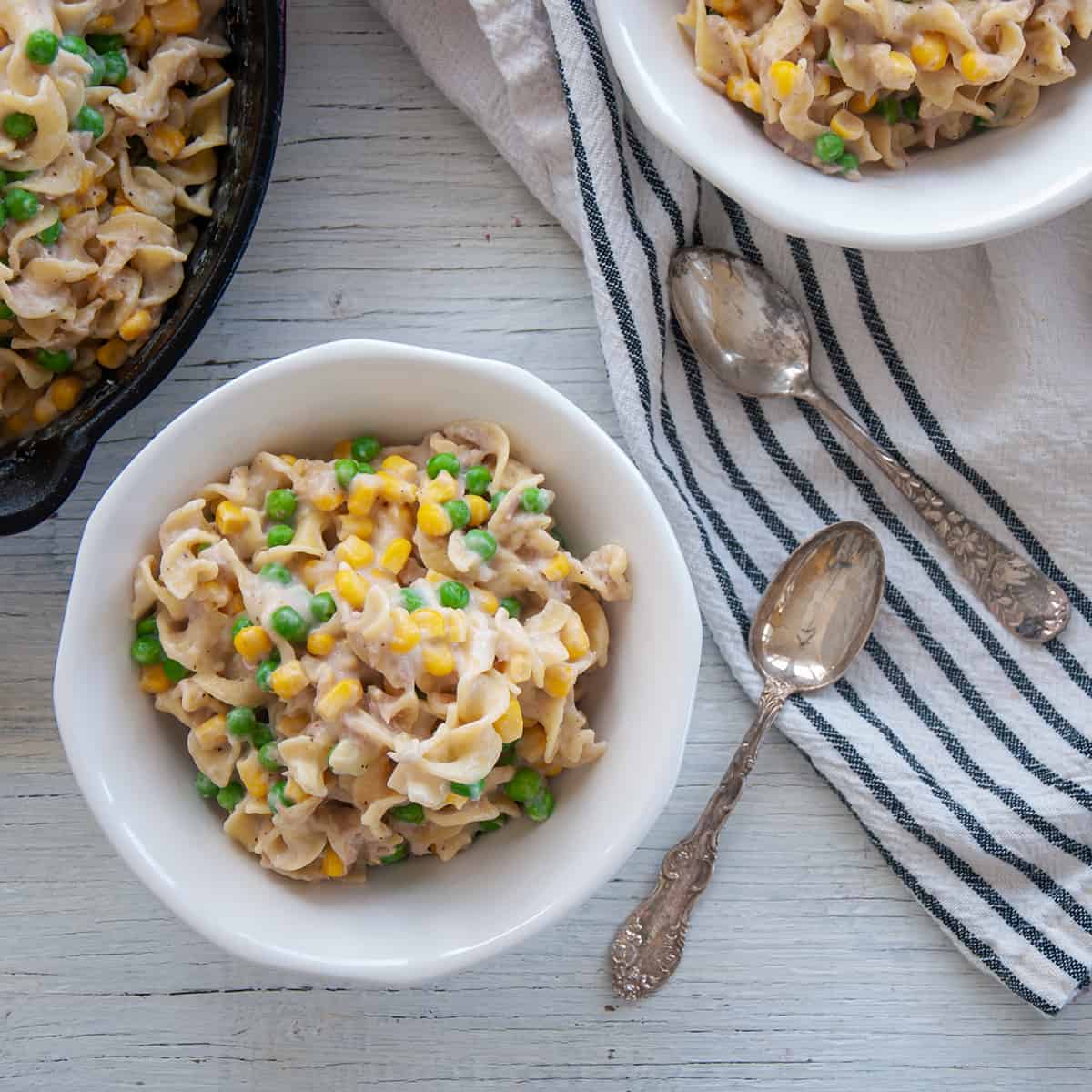 Two white bowls filled with Homemade Skillet Tuna Helper and two silver spoons on a white and blue cloth.