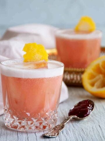 Two Strawberry Jelly and Gin Cocktails on a white table with a spoon of jelly and a juiced lemon on the side.