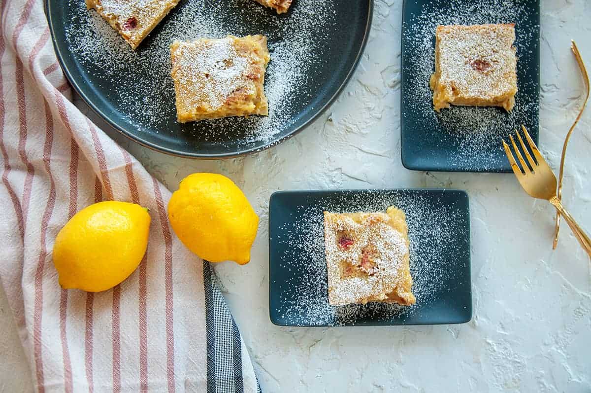 Overhead view of three Lemon Rhubarb Bars on dark plates and sprinkled with powdered sugar. There are a few whole lemons and two gold forks next to the plates.