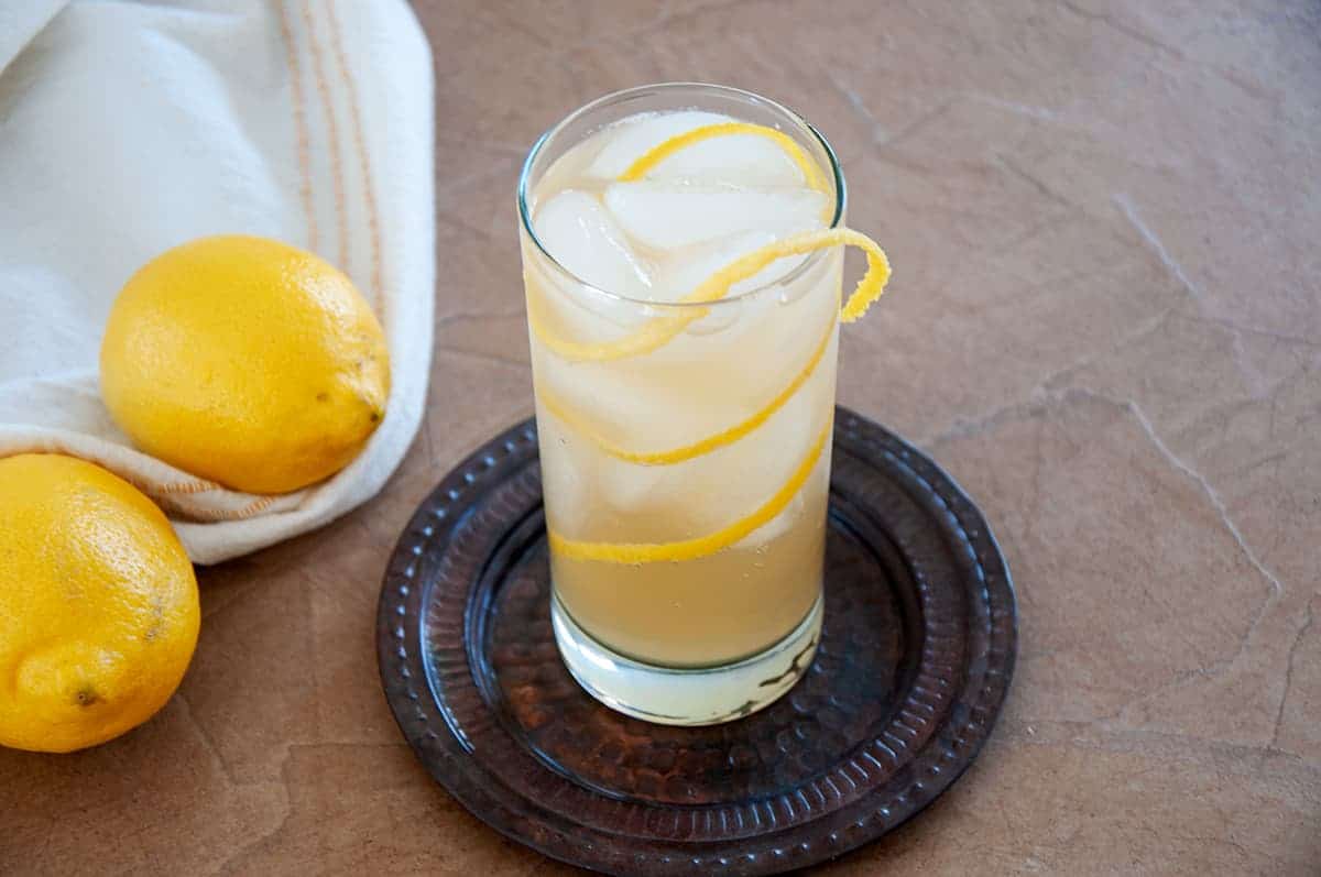 One full Horse's Neck cocktail on a brown plate and on a textured table with two whole lemons showing to the side.