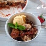One serving of Strawberry Rhubarb Cobbler with a side of vanilla ice cream and garnished with a few leaves of mint. This is served in a white bowl with a silver spoon.