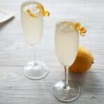 Two French 76 Cocktails on a white table with lemon peel garnish and a whole lemon in the background.