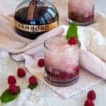 Two Raspberry Gin Smash Cocktails, garnished with mint, on a white table that has raspberries laid across it and a bottle of Chambord.