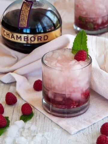 Two Raspberry Gin Smash Cocktails, garnished with mint, on a white table that has raspberries laid across it and a bottle of Chambord.