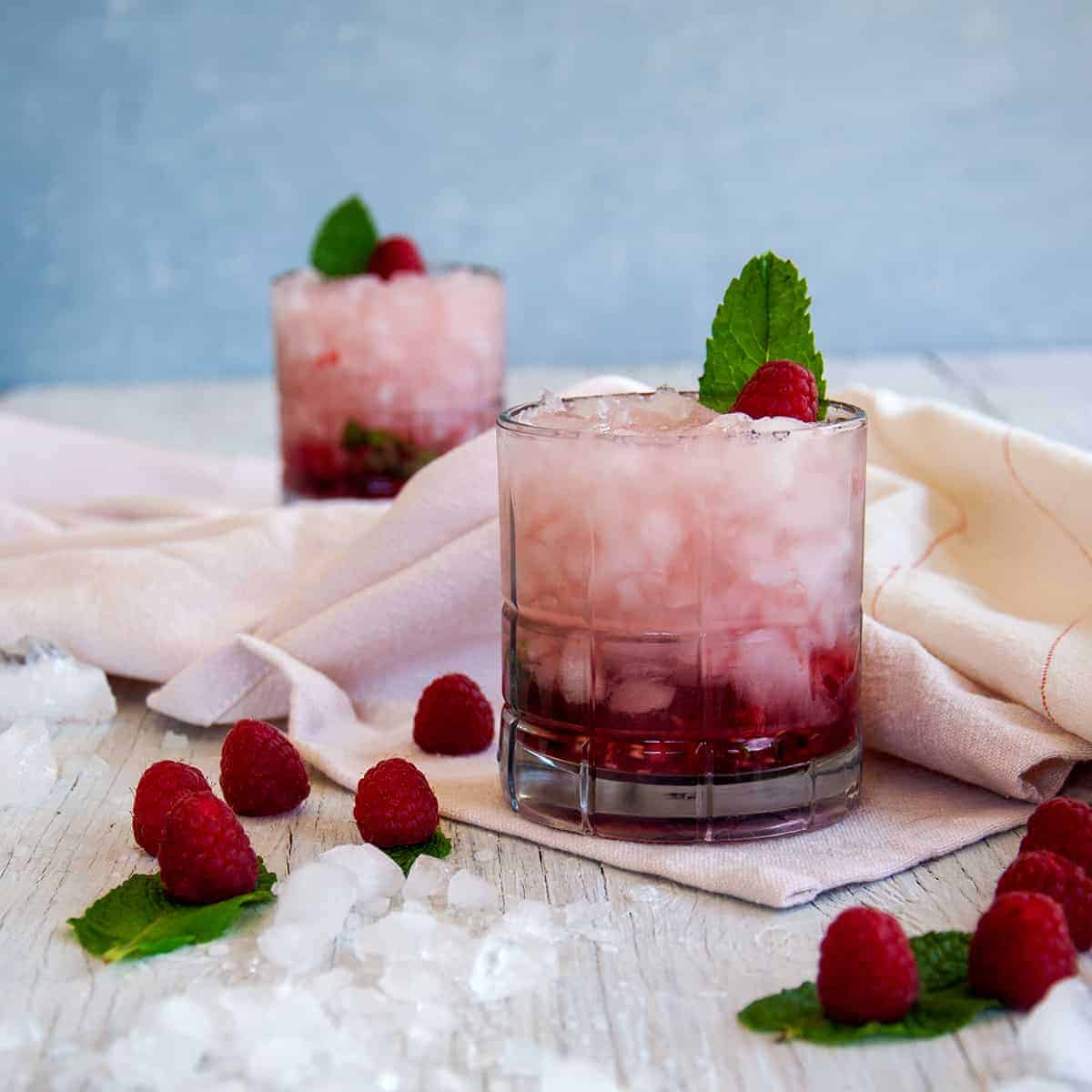 Two Raspberry Gin Smash Cocktails, garnished with mint, on a white table that has raspberries laid across it.