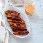 Plate of bacon-wrapped grilled sweet potato fries on a white plate with a mason jar of sriracha maple dipping sauce to the side.