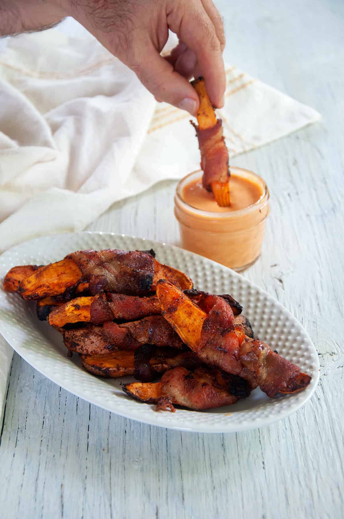 Plate of bacon-wrapped grilled sweet potato fries with a mason jar of sriracha maple dipping sauce to the side and a hand dipping one fry into it.