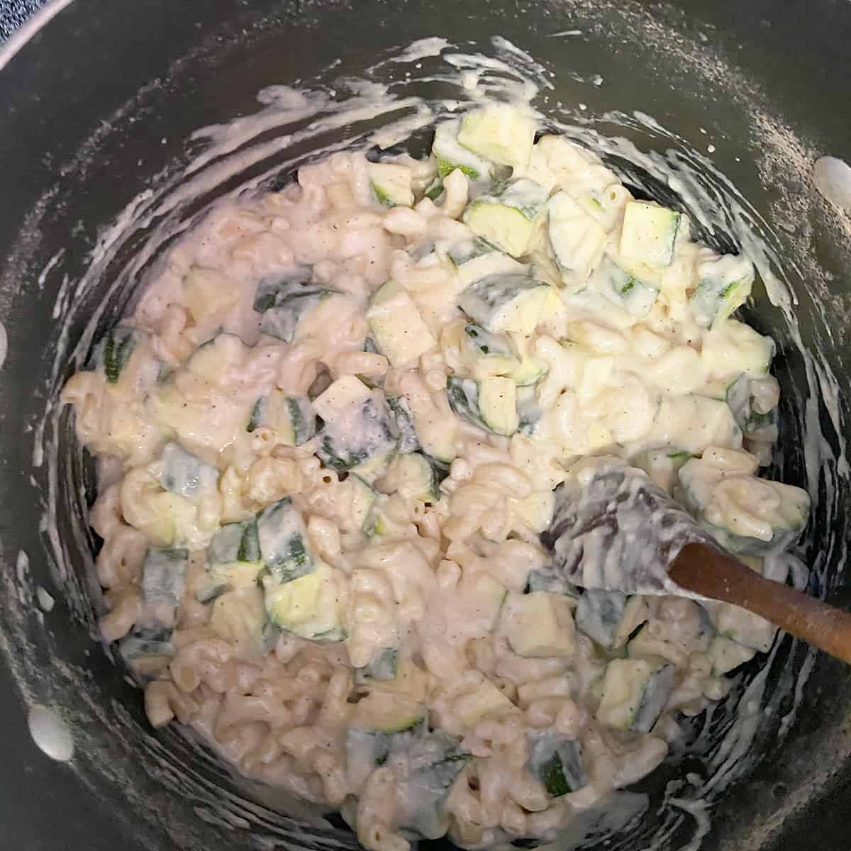 Dish where zucchini, macaroni, and cheese sauce are being mixed with a wooden spoon.