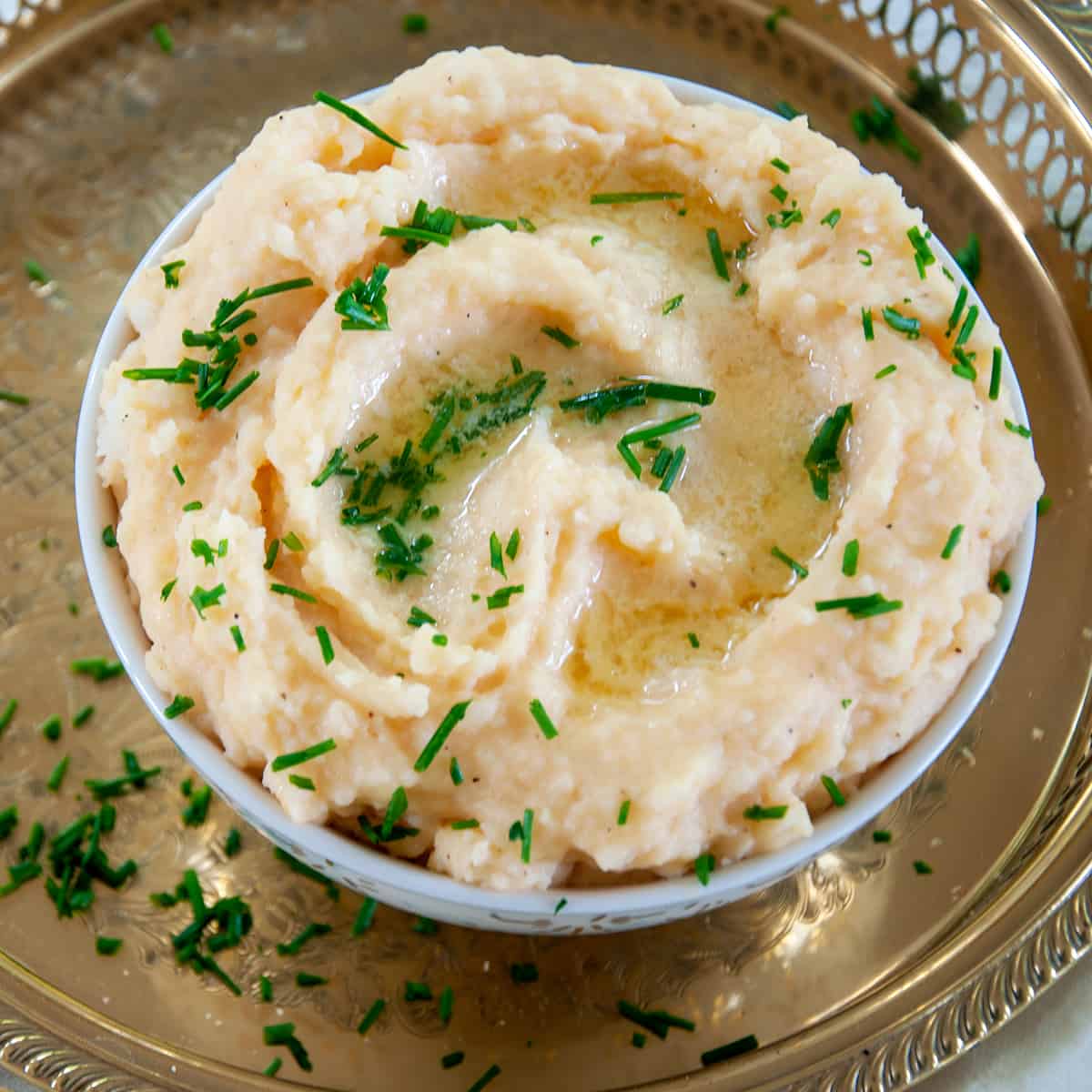 Bowl of Cheesy Mashed Potatoes in a white bowl that is sitting on a platter and chopped chive sprinkled all over.