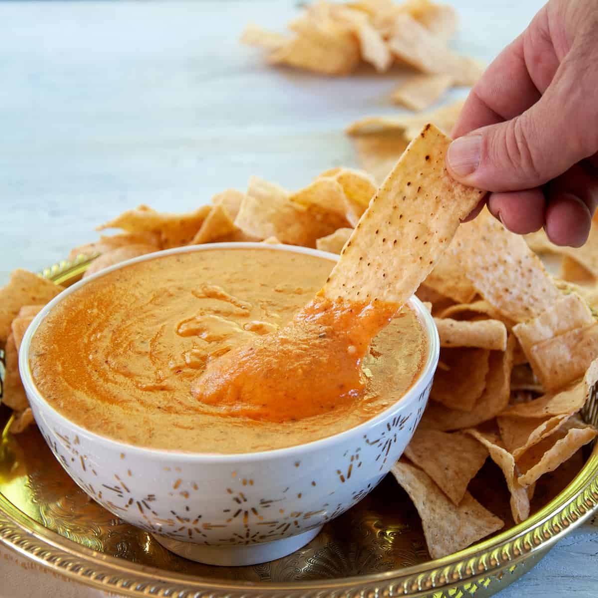 Homemade Queso Dip in a white and gold bowl and a hand dipping a single corn chip into it.