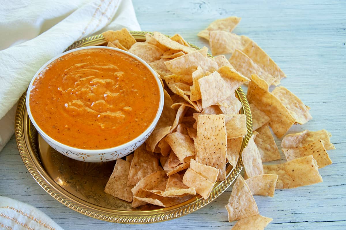 Bowl of queso dip on a gold platter with corn chips spread all around the platter and the table underneath.