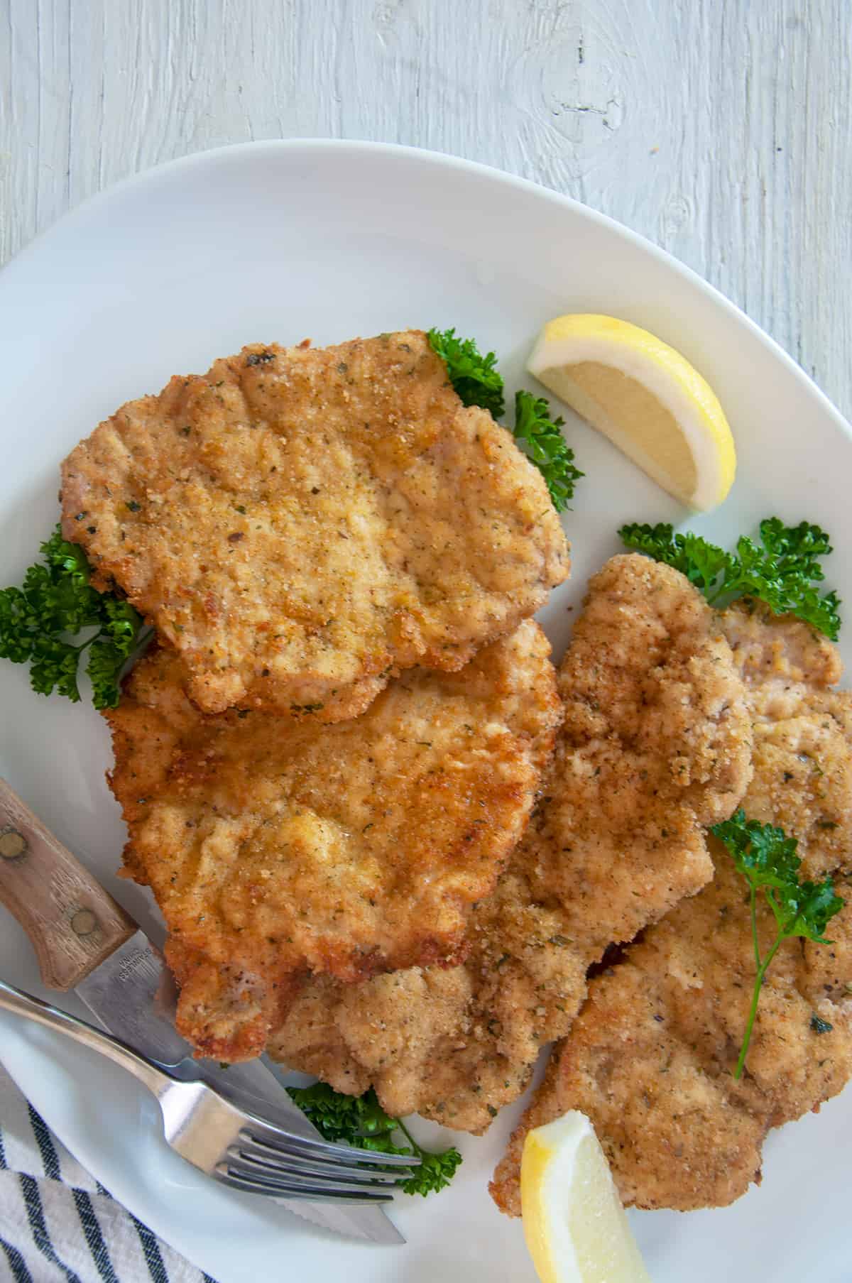 Overhead view of four pork schnitzel cutlets on a white platter, with a fork and knife off to the side.