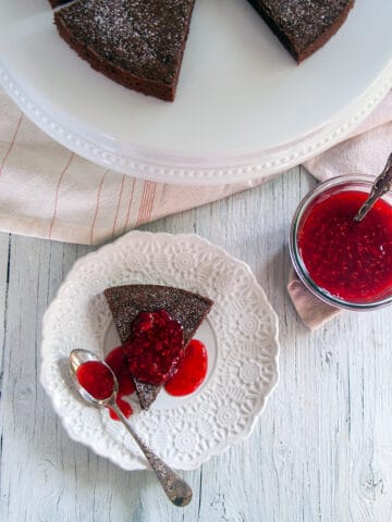 One slice of our Kladdkaka Swedish Sticky Cake on a white plate, covered in raspberry topping and a spoon to the side.