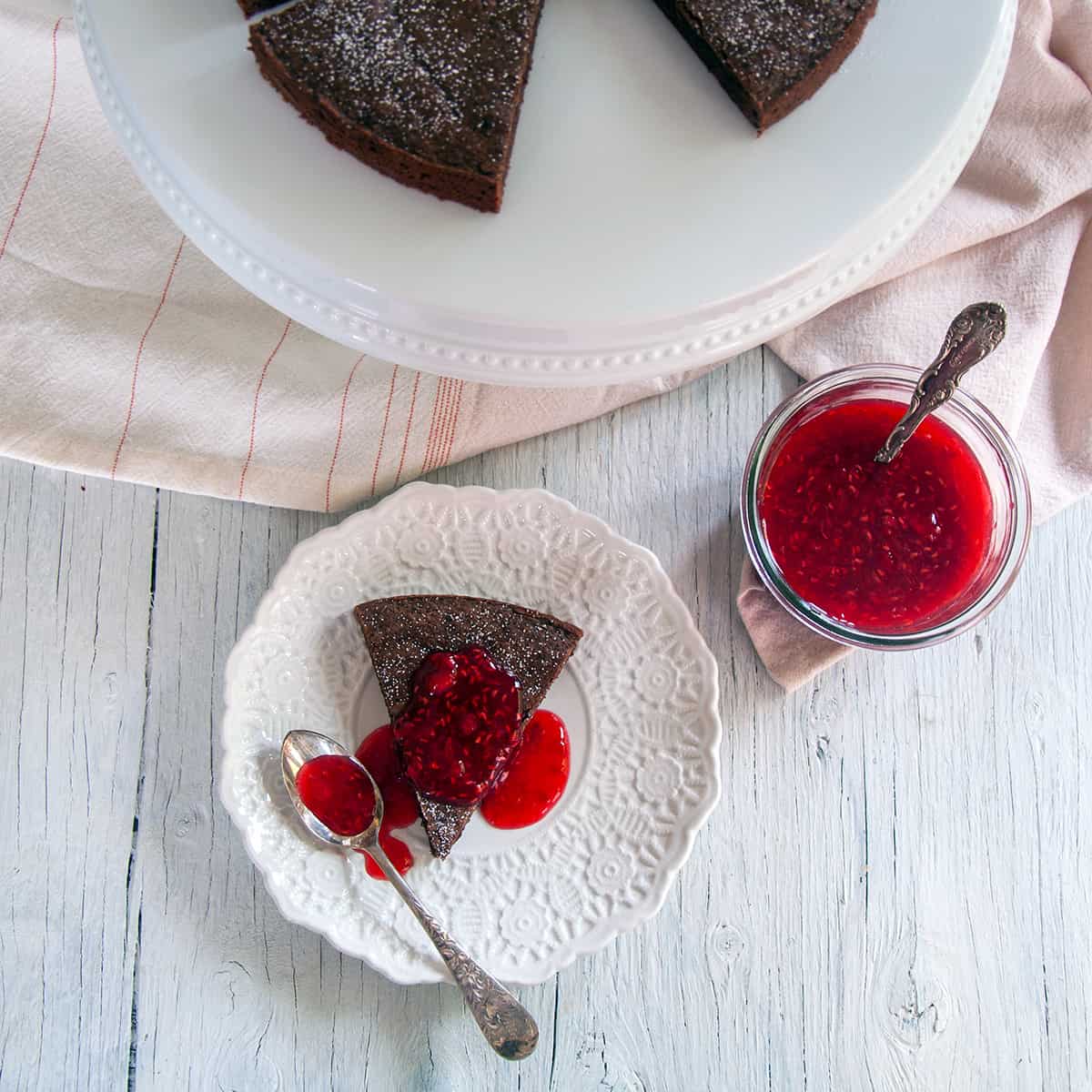 One slice of our Kladdkaka Swedish Sticky Cake on a white plate, covered in raspberry topping and a spoon to the side.