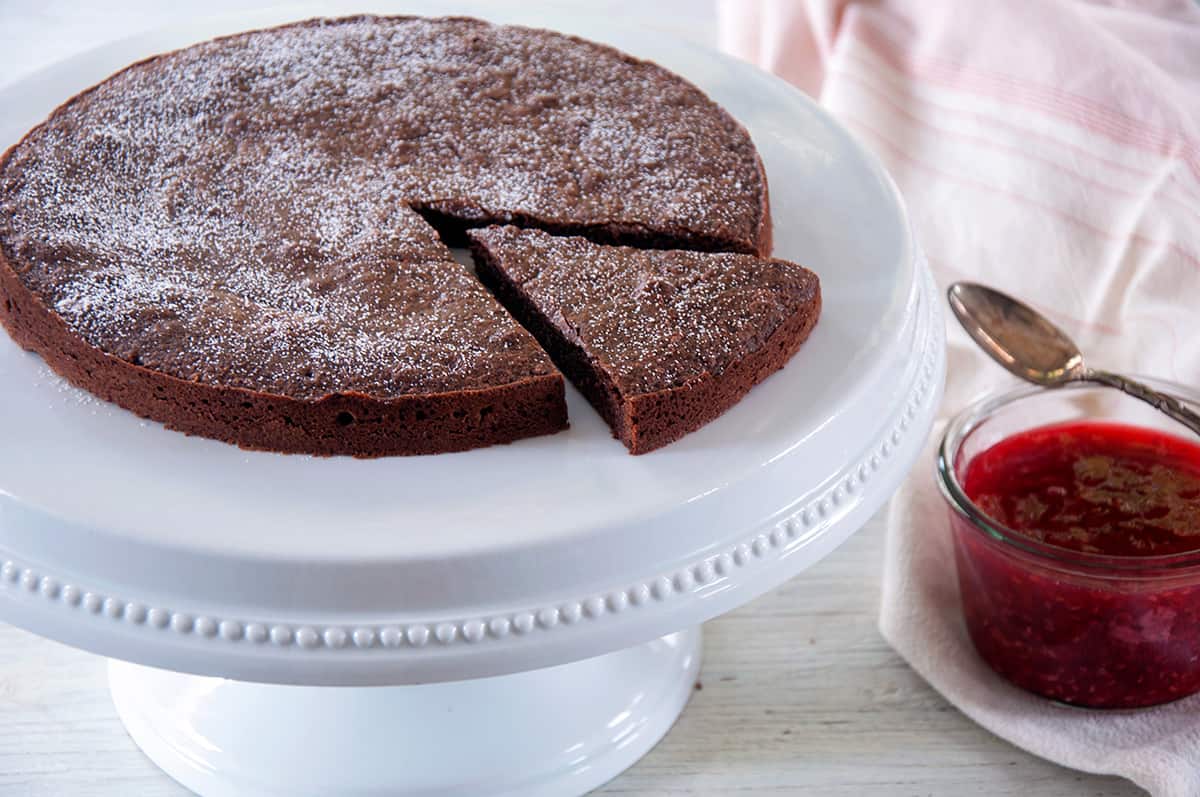 The entire Kladdkaka on a white serving platter with one slice cut out and a bowl of the raspberry topping off to the side.