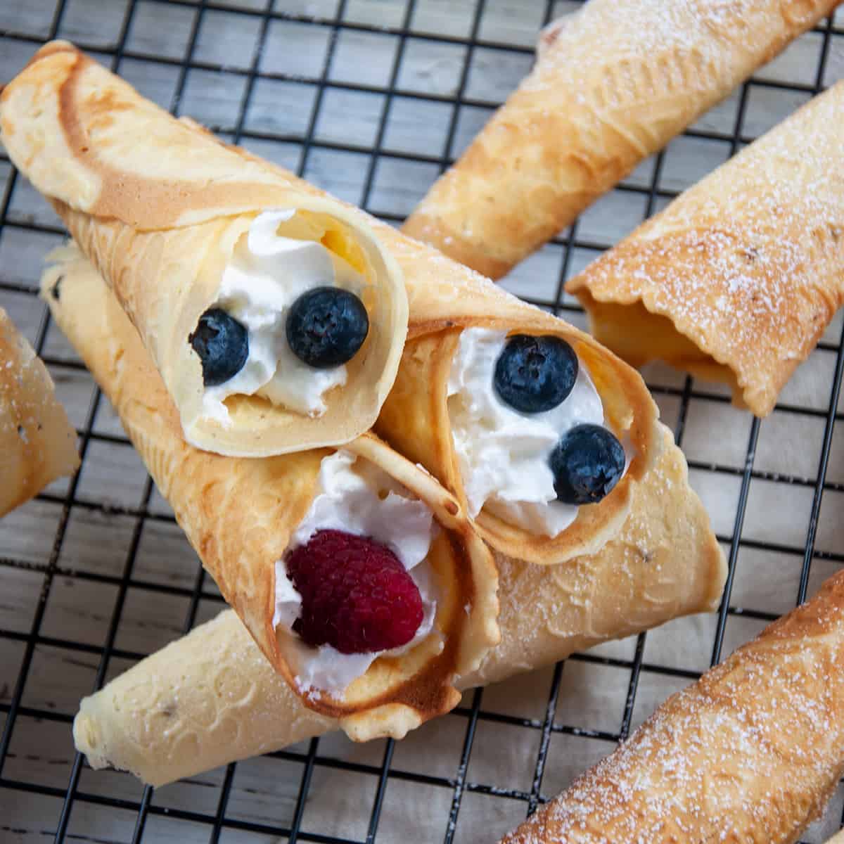 Three Krumkake filled with whipped cream, blueberries, and raspberries that are laying on top of several unfilled ones that have been sprinkled with powdered sugar.