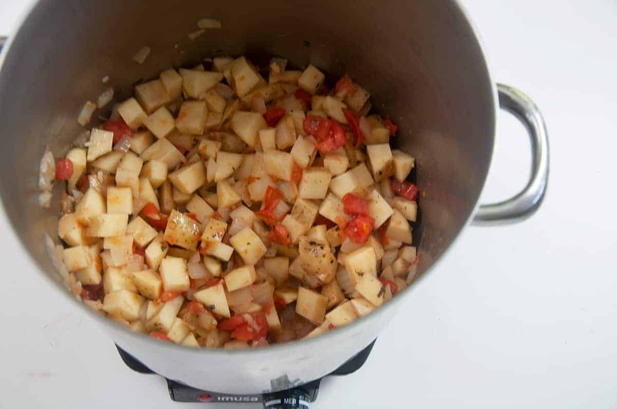 Pot of potato, onion, tomato, and oregano cooking over medium heat on a white table.