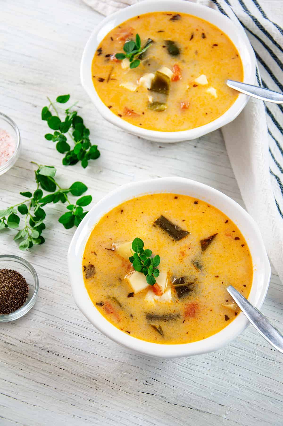 Two bowls of Caldo Queso on a white table with salt, pepper, and oregano.