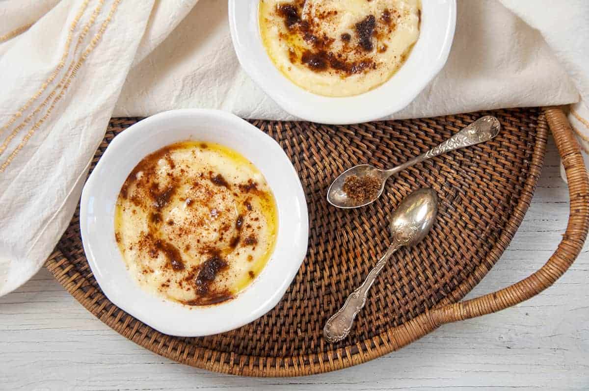 Two bowls of Rømmegrøt that are on a wicker platter with a few antique spoons to the side.