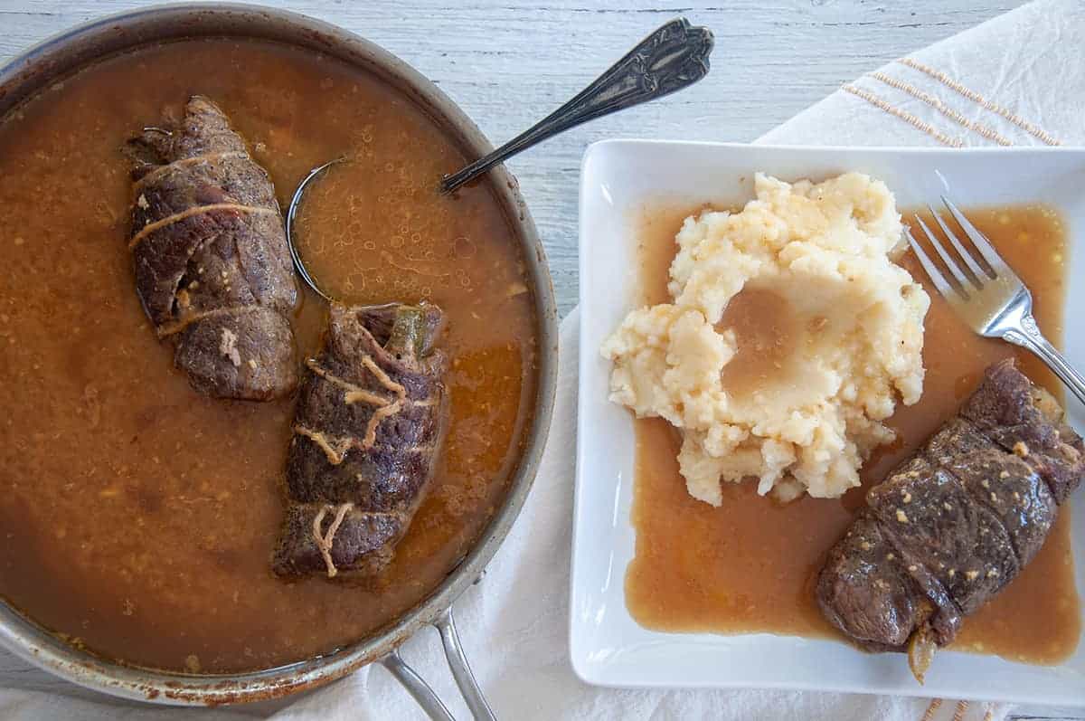 One rouladen on on a white plate with gravy and mashed potatoes, while two others are still sitting in the simmering pan.
