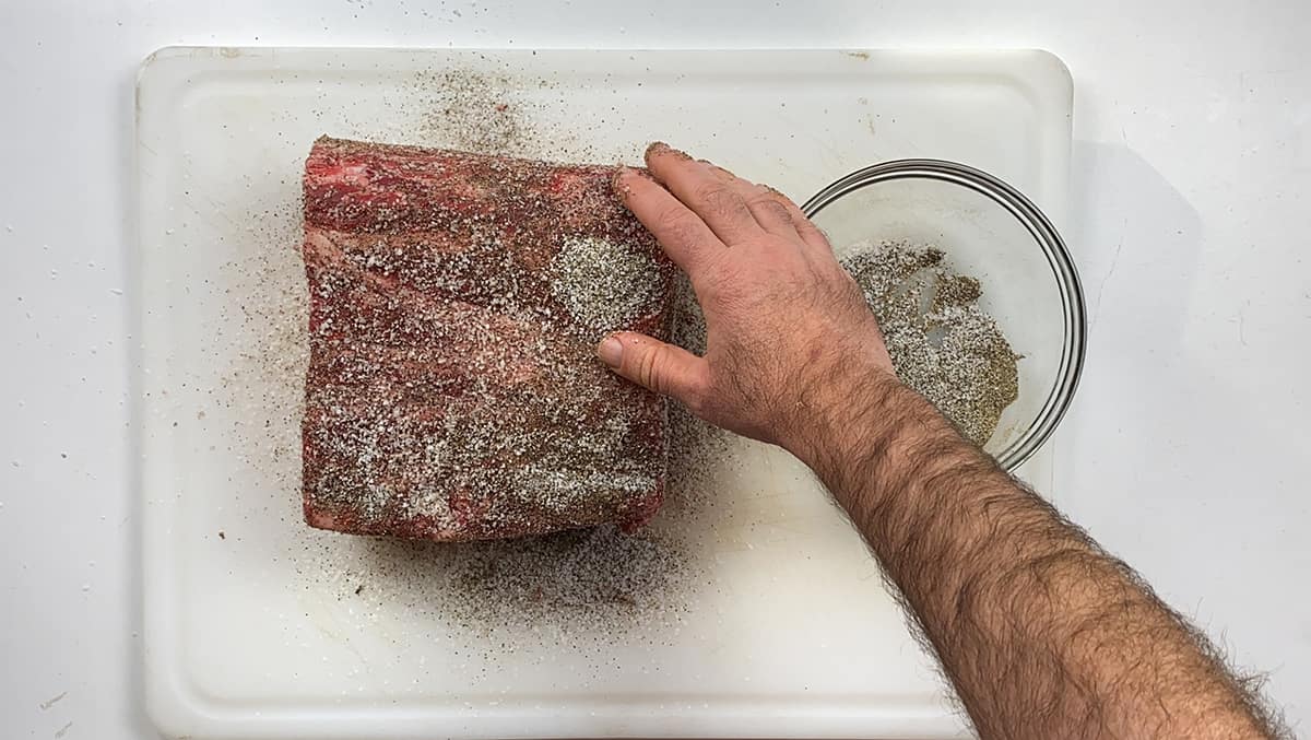 A raw prime rib on a cutting board with a salt & pepper mixture being rubbed all over it.