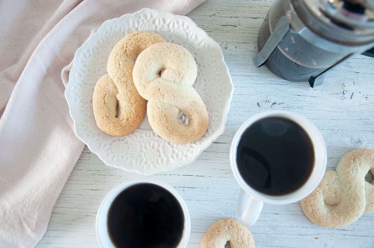 Two kringler on a white plate with two cups of coffee and a French press off to the side.