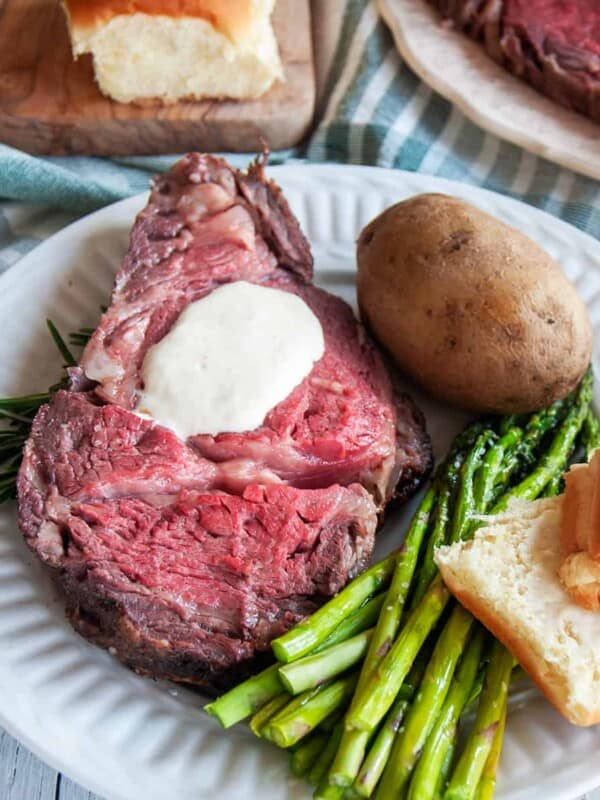 Slice of Prime Rib on a white plate being served with asparagus, a bun, a baked potato, and the entire roast barely showing in the background.