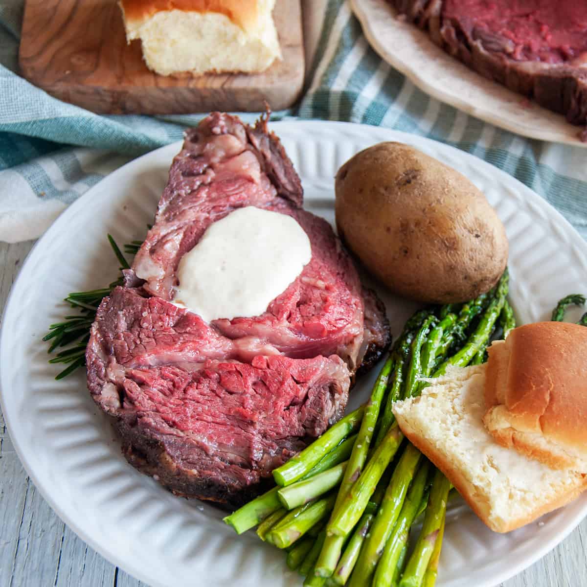 Slice of Prime Rib on a white plate being served with asparagus, a bun, a baked potato, and the entire roast barely showing in the background.