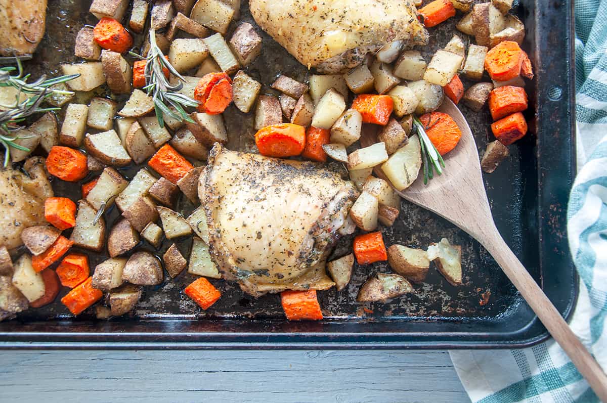 Four baked italian chicken thighs on a baking pan with potatoes and carrots.