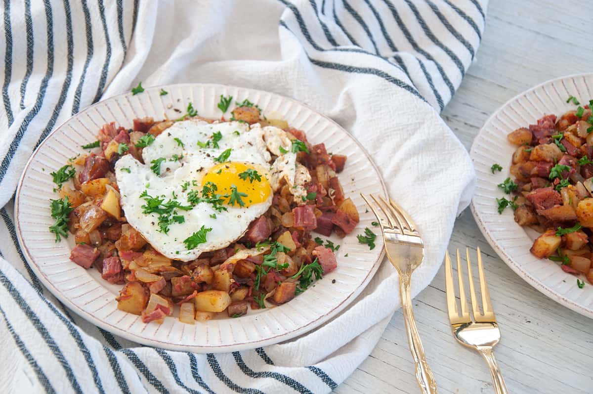 Corned Beef Hash covered with an egg, garnished with parsley, and two gold forks to the side.