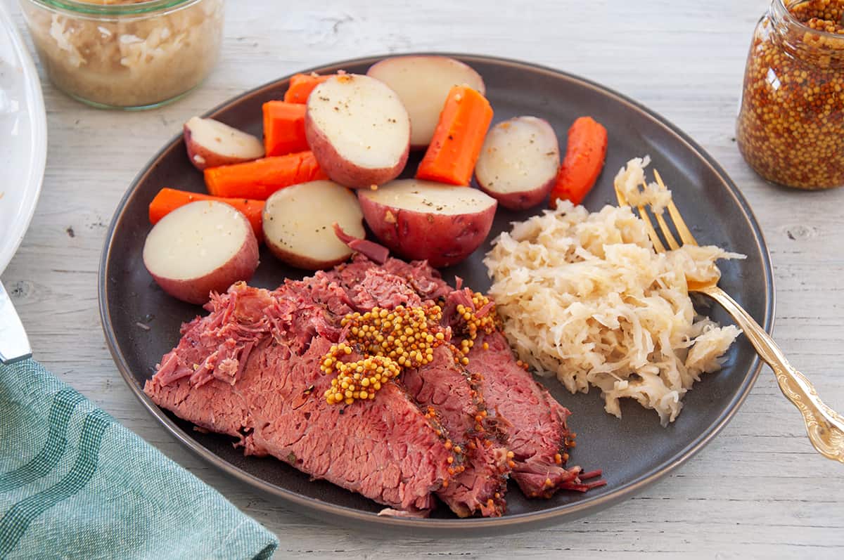 Corned Beef cuts, covered with whole grain mustard, and potatoes, carrots, and sauerkraut on the side of a plate.