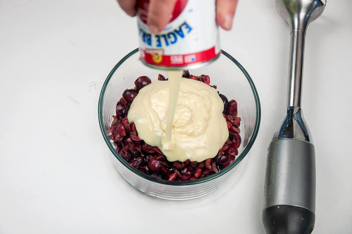 Pouring sweetened condensed milk into a bowl of chopped cherries.