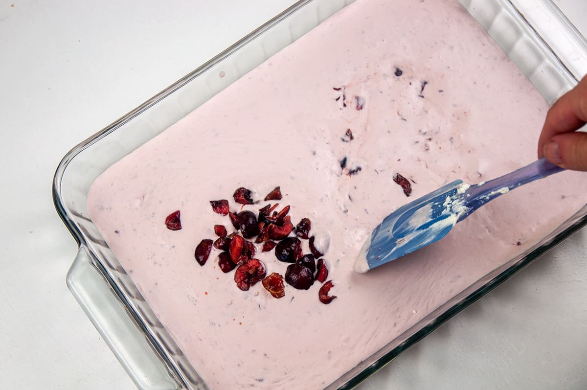 A 9x13 baking dish filled with unfrozen ice cream and a blue spatula mixing in chopped cherries.