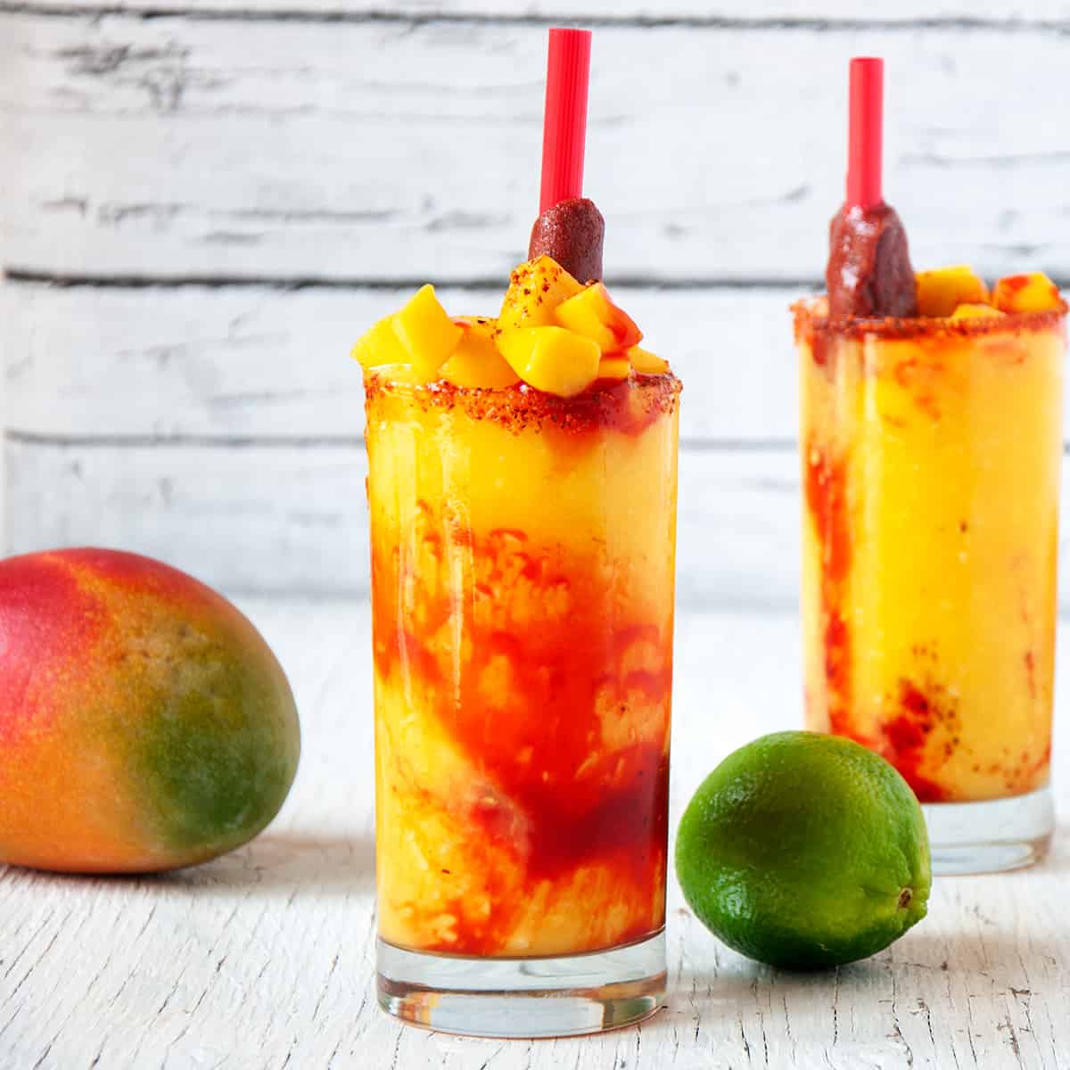 Two Mangonadas with Tamarind candy straws on a white table with a mango and lime.