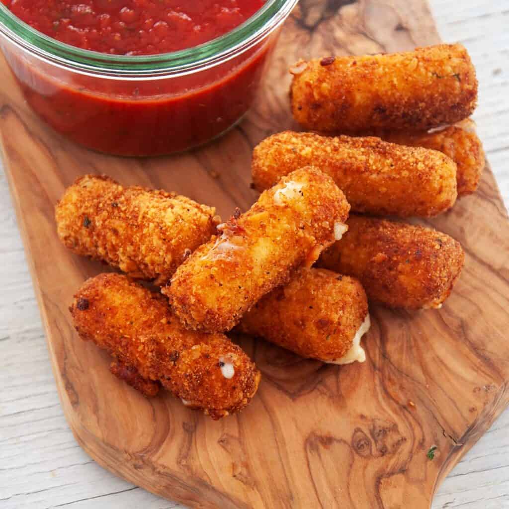 A stack of fried cheese sticks on a wood platter and a side of marinara sauce.
