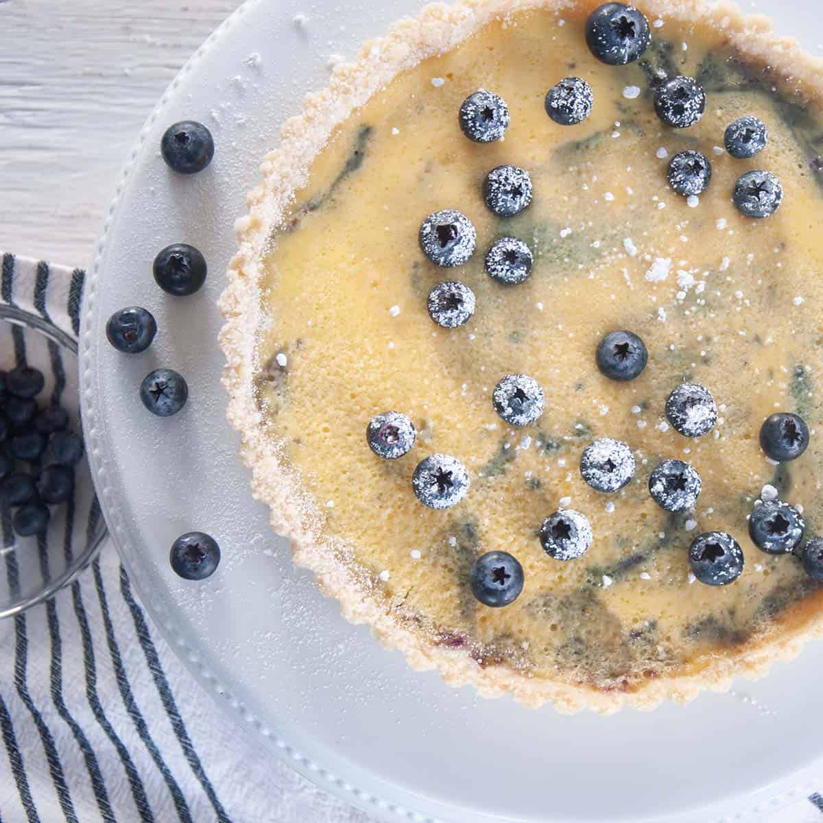 Whole blueberry tart with fresh blueberries on top and a on a white platter.