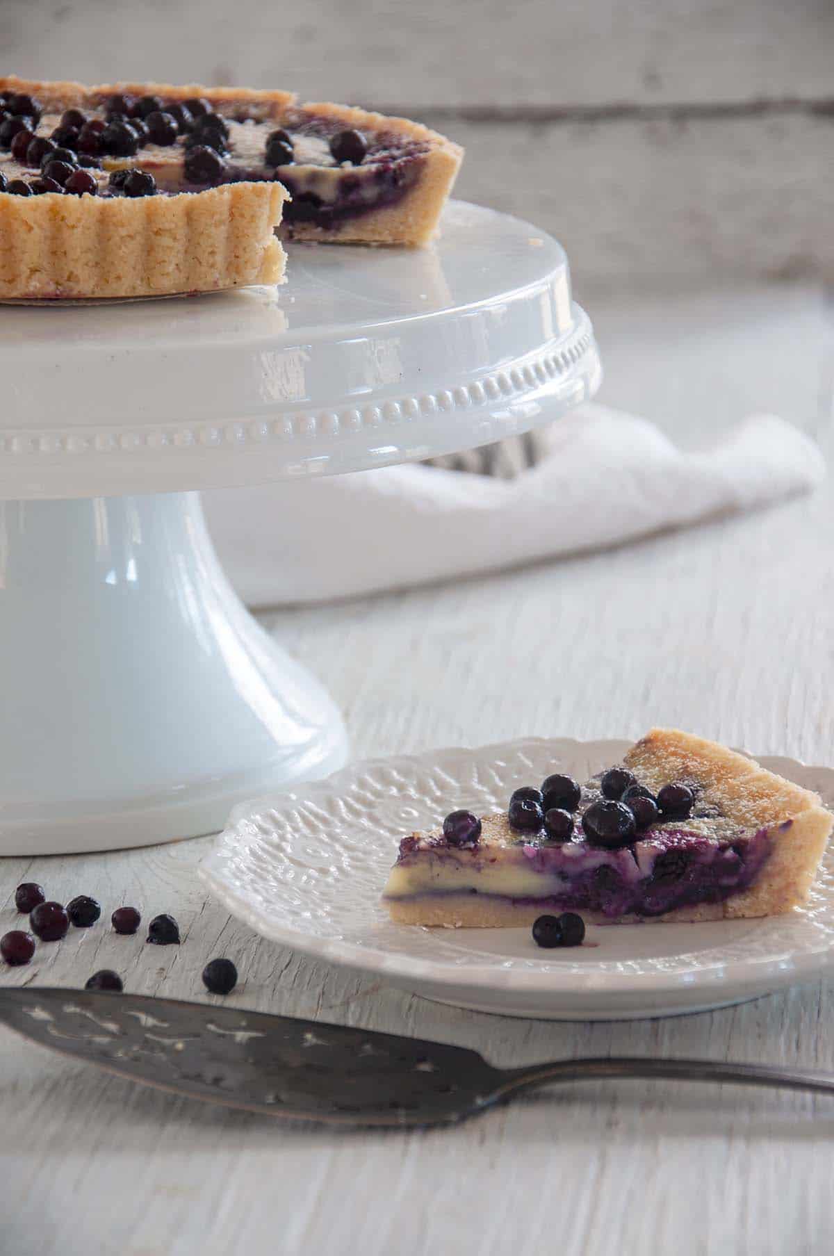 Single slice of blueberry tart on a white plate with the rest of the tart on a platter.