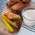 Cheesy Fried Pickle cut in half on a cutting board with full egg rolls and dip.