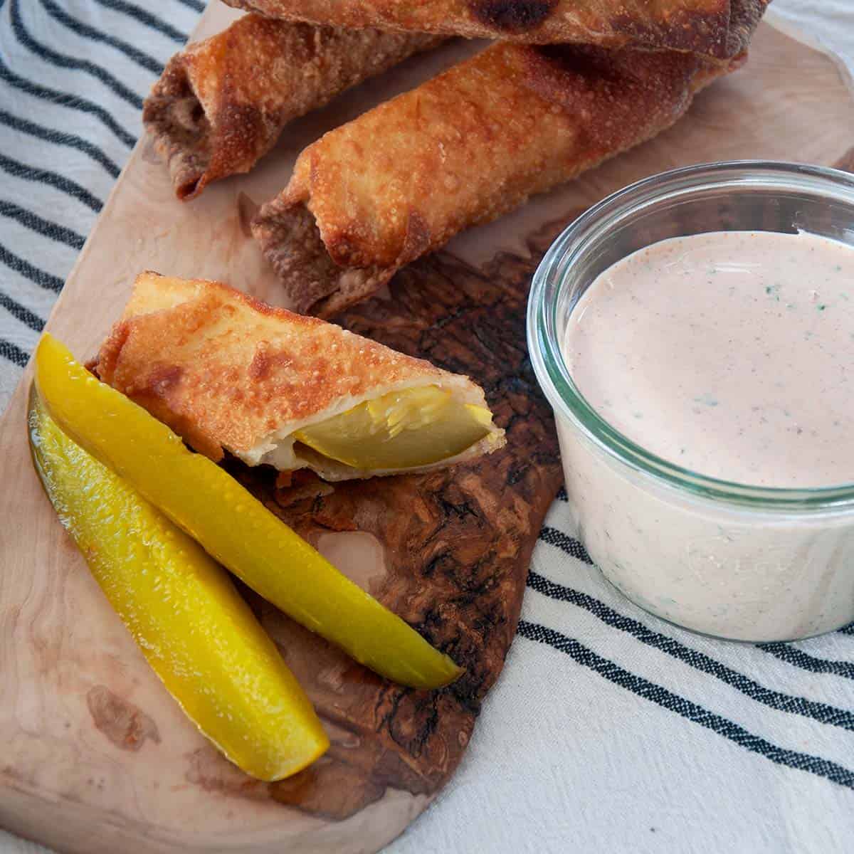 Cheesy Fried Pickle cut in half on a cutting board with full egg rolls and dip.