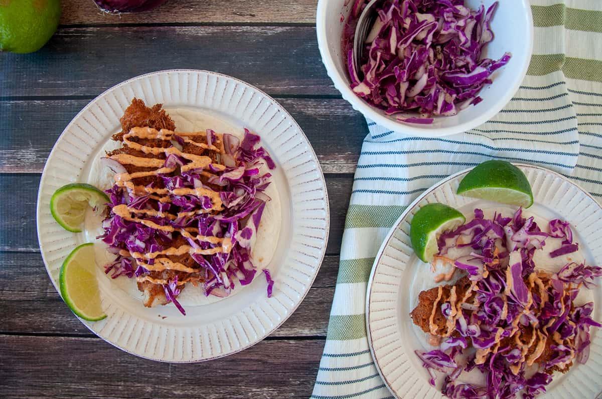 Two fish tacos on white plates with limes to the side.